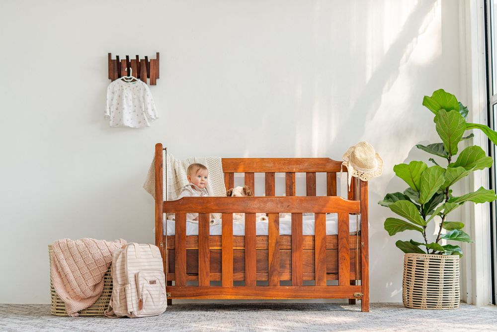 Baby sitting in a wooden cherry crib in a bright nursery with a fiddle leaf fig plant and wicker basket nearby.