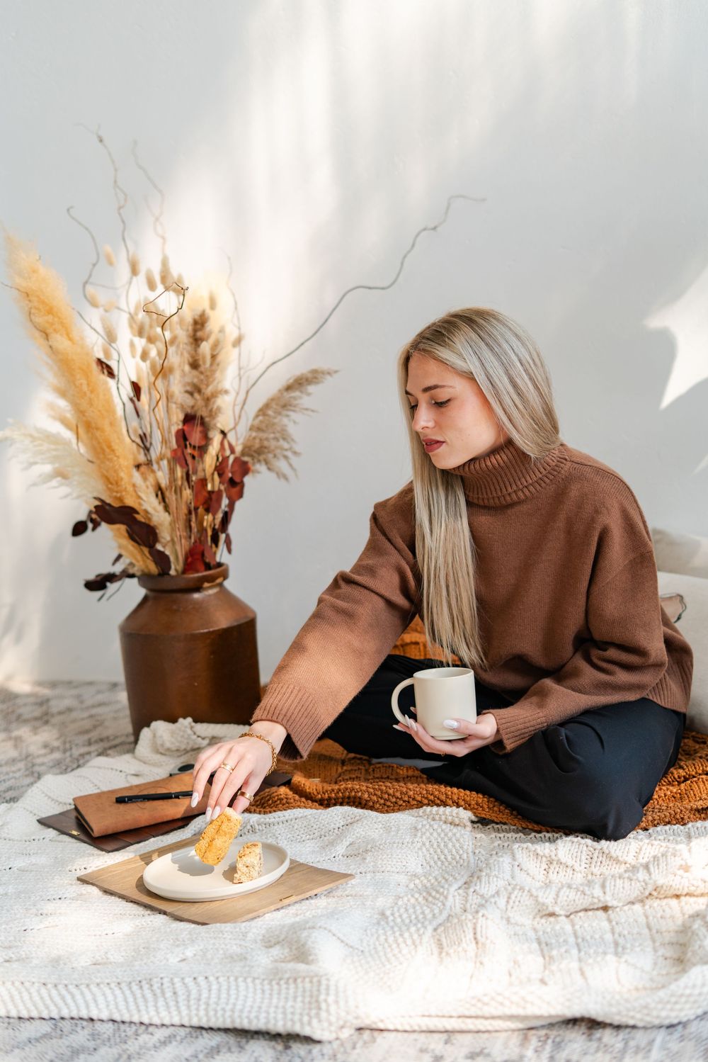 Blonde woman photographed in brown turtleneck sweater sitting on floor with coffee cup, reaching for pastry near dried floral arrangement.