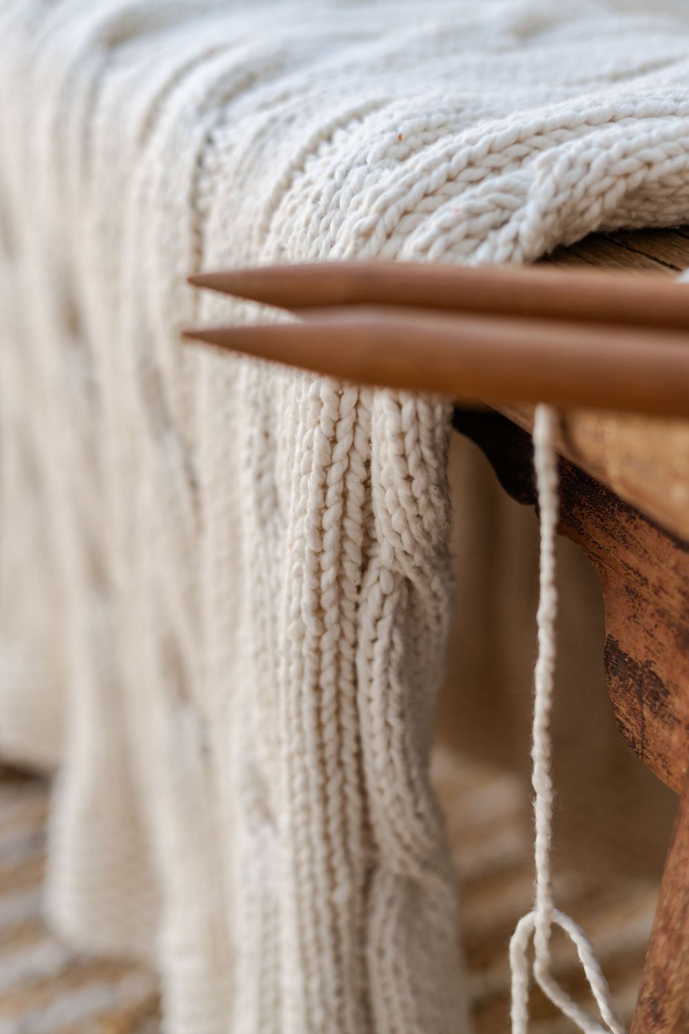 Close-up of cream cable-knit blanket draped over wooden furniture with brown knitting needles and yarn during a product shoot.