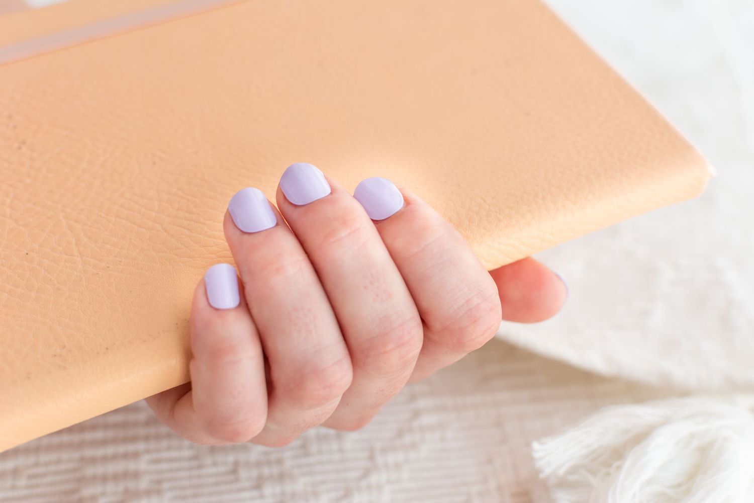 Close-up of fingers with light purple nail polish on a peach-colored background.