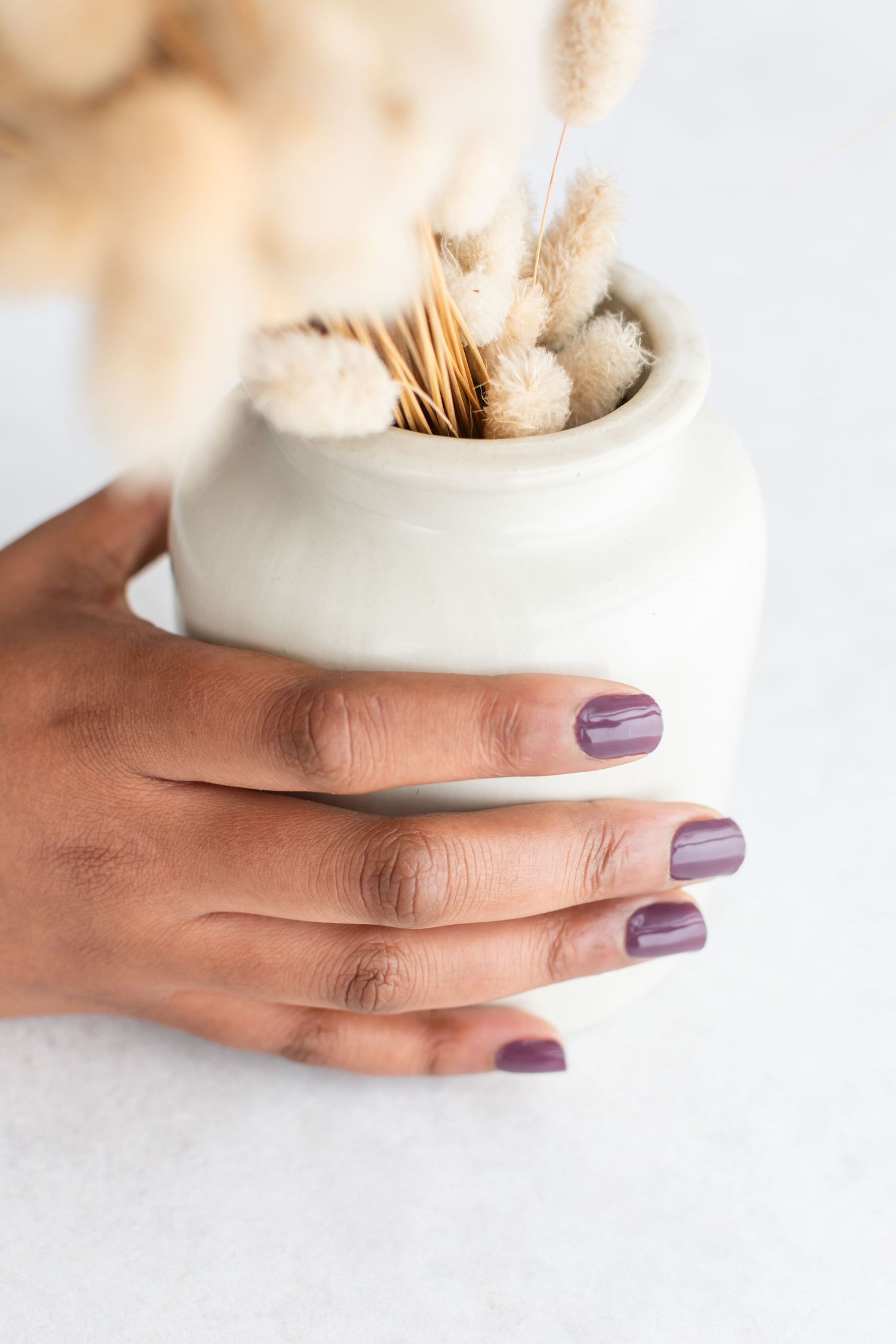 Hand with purple manicured nails holding a white ceramic container filled with dried flowers and cotton stems.