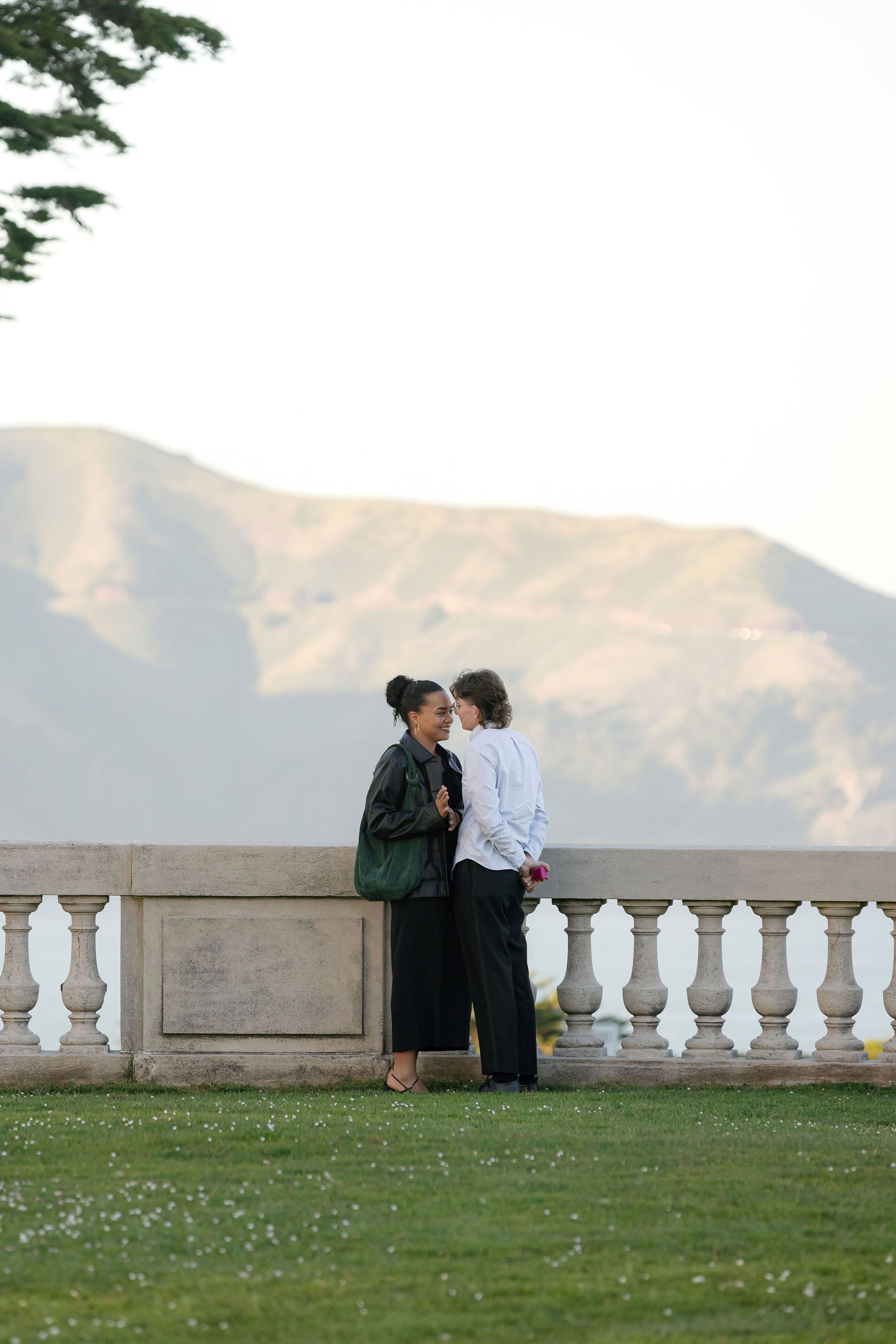 A romantic couple shares an intimate moment on a terrace with a mountainous backdrop and white stone balustrade.