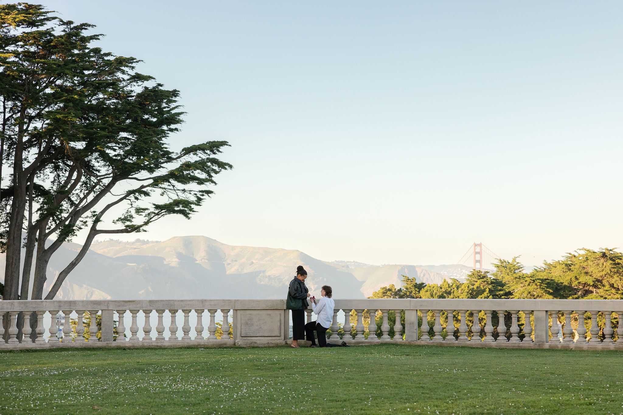 A silhouetted couple stands on a scenic terrace with ornate white railings overlooking a sunset-lit landscape.