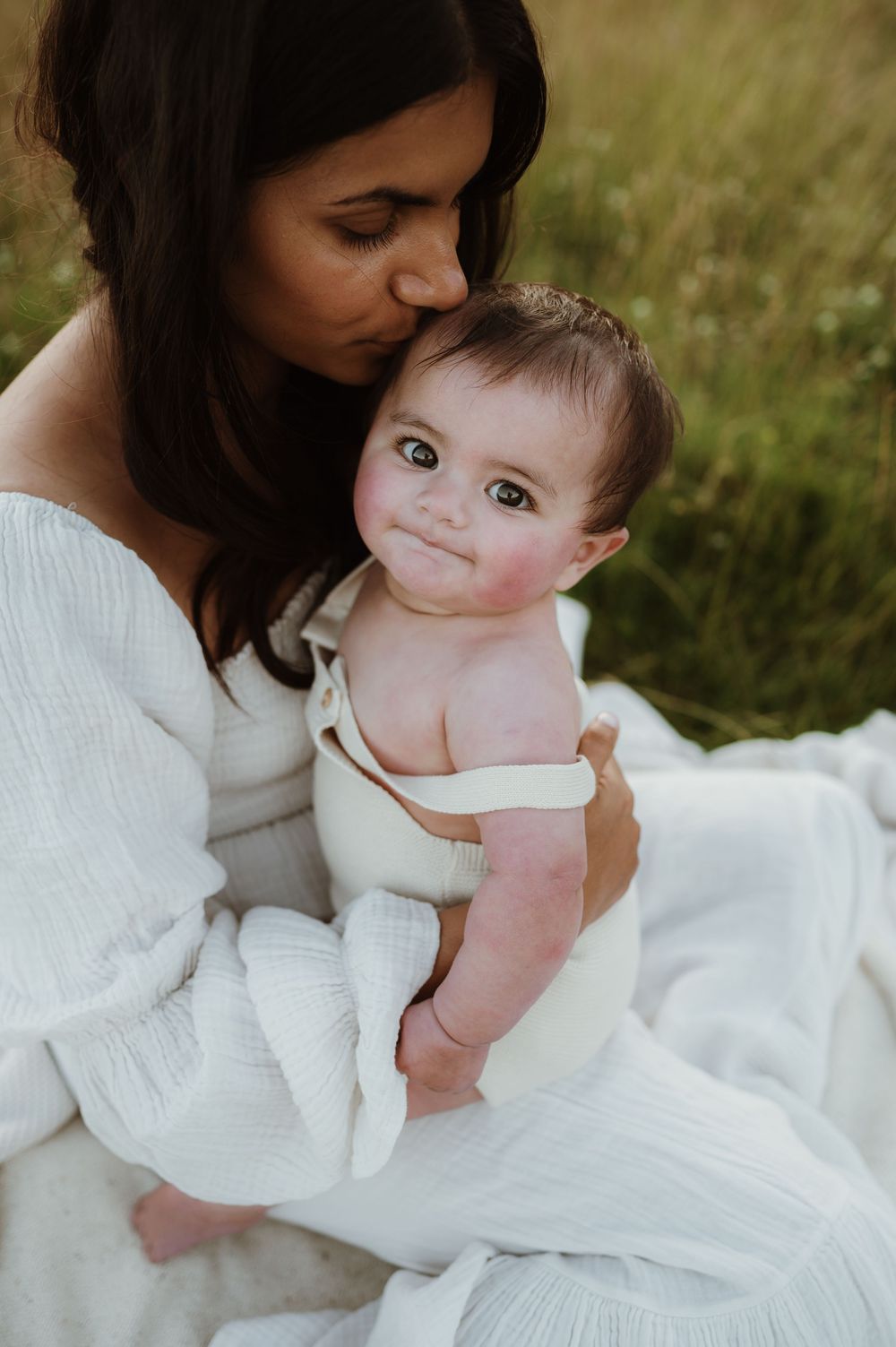 An intimate moment captured as a mother in a white dress holds her baby outdoors in a grassy field.