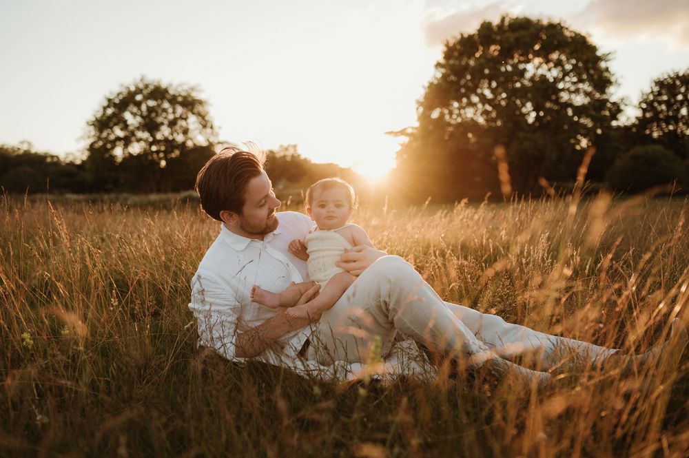 A parent and child share a tender moment in a sunlit wheat field at golden hour while sitting among tall grass.
