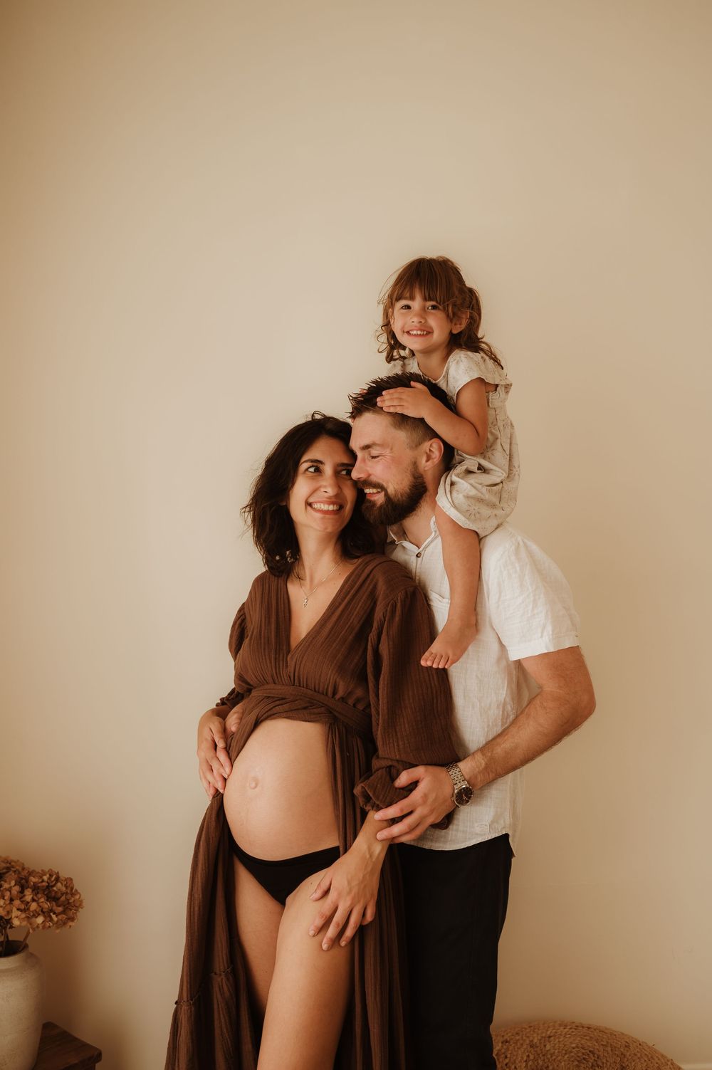 A couple in brown and white outfits share tender moments during maternity photo session against neutral wall.