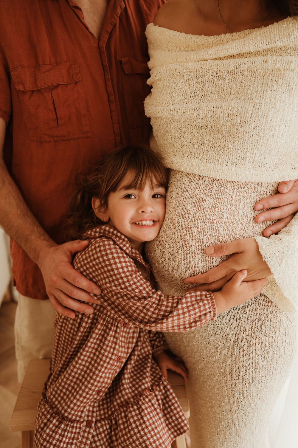 A small child wearing a checkered dress embraces a pregnant person in a cream dress while standing next to a figure in rust.