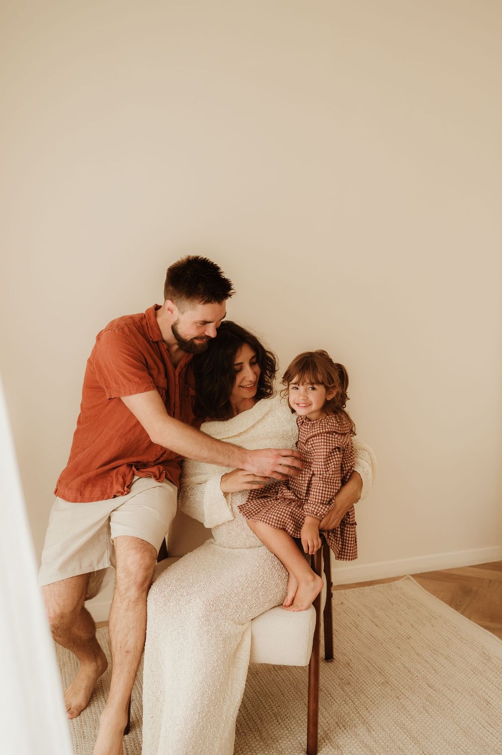 A cozy family moment shows a group sitting together on a chair in warm earth tones against a beige wall.