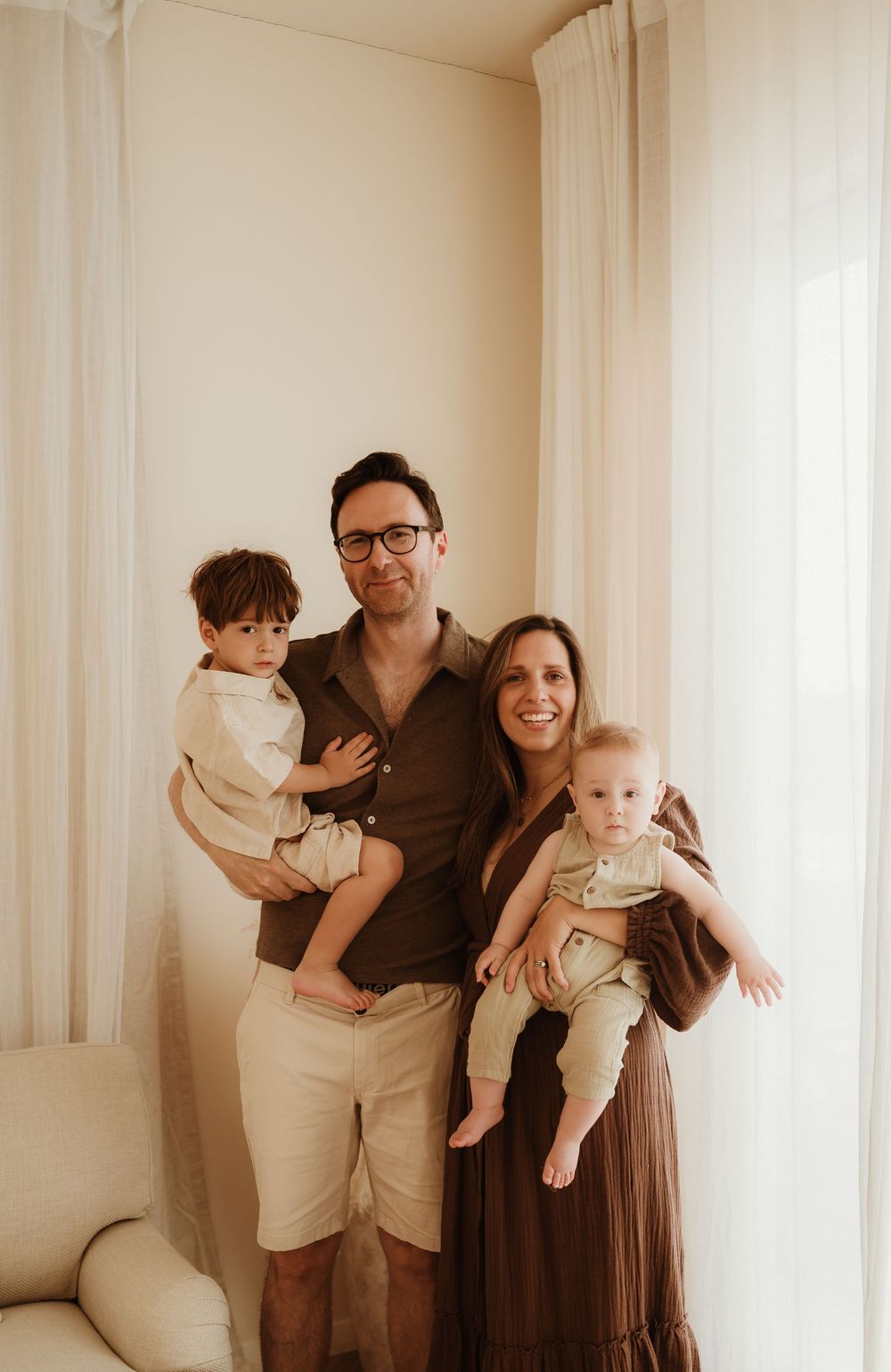 A family in neutral tones poses together in a casual indoor setting with light curtains.
