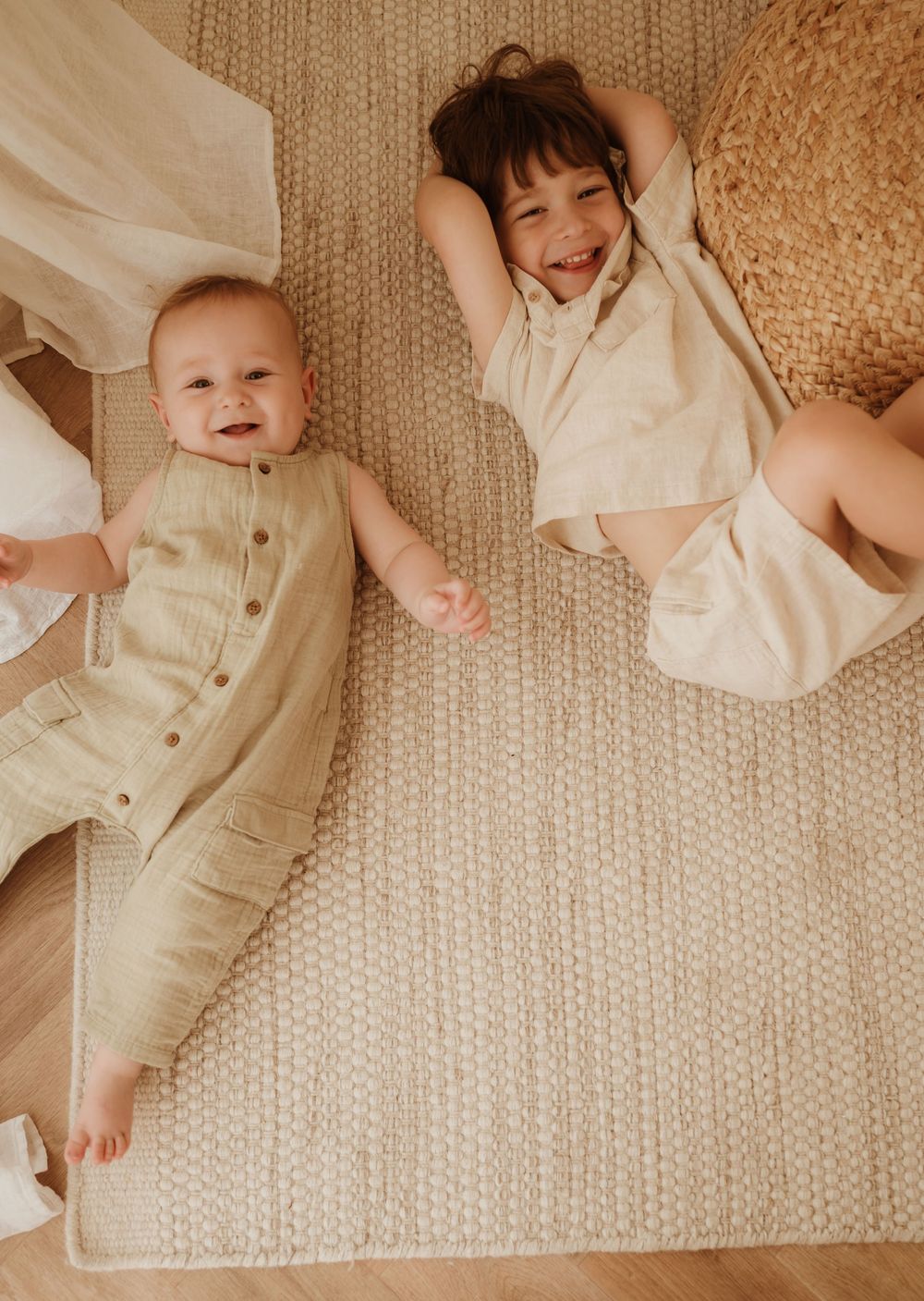 Baby wearing cream-colored romper lies on knitted blanket with light fabric draping in multiple artful angles.