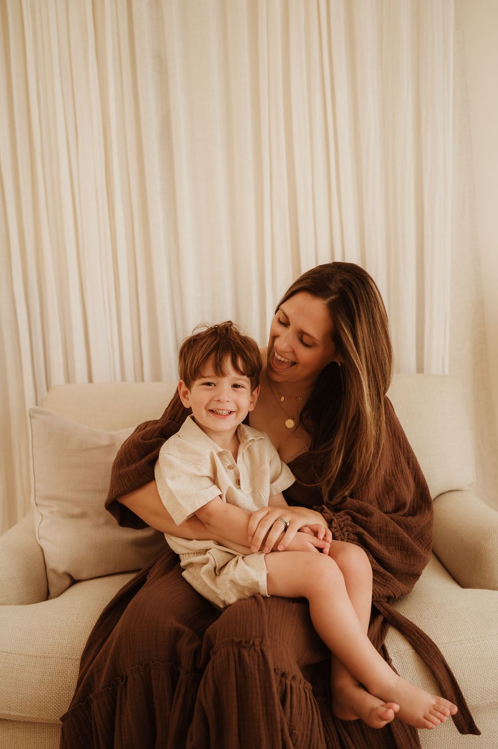 A tender moment shared between parent and child cuddling on a cream colored couch with white curtains in background.