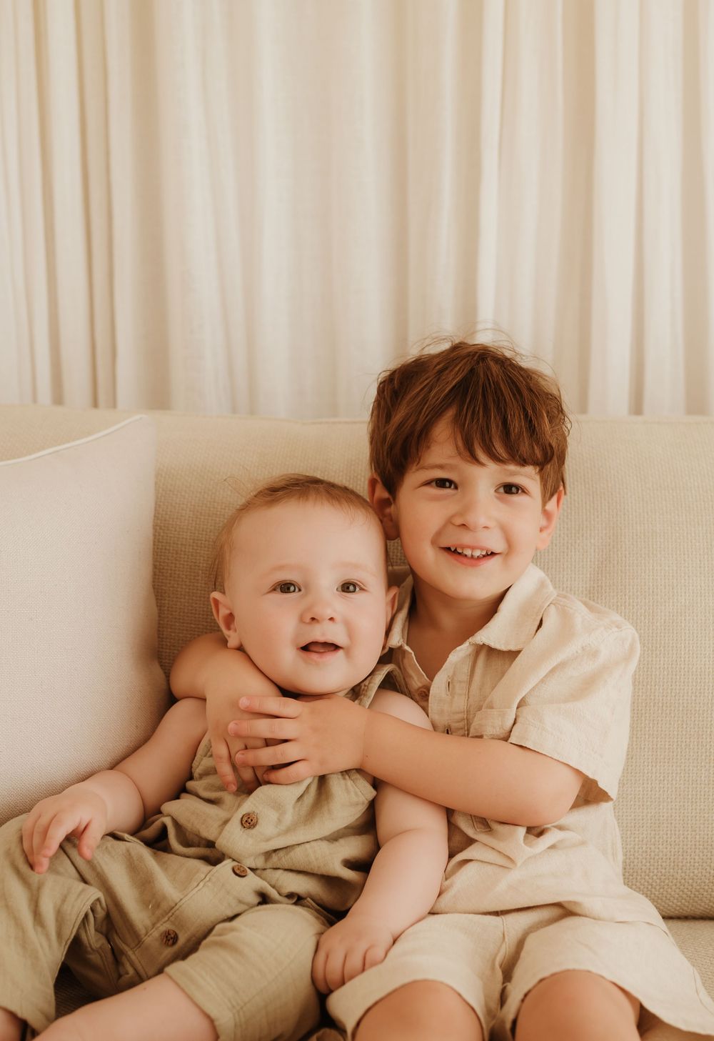 Two young children in beige outfits sitting together on a cream-colored couch against a light curtained backdrop.