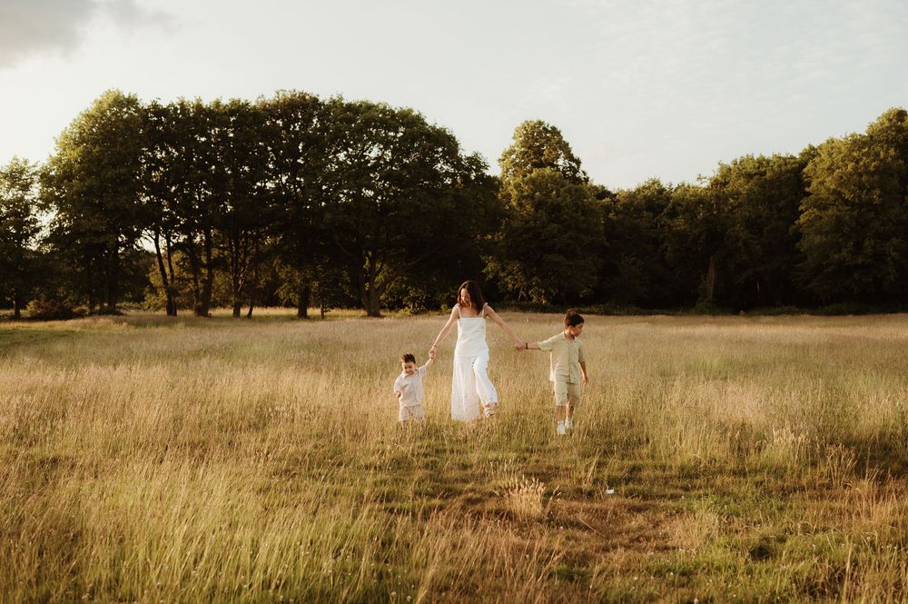 Three people in white dresses walk through a golden field at sunset with trees in the background.