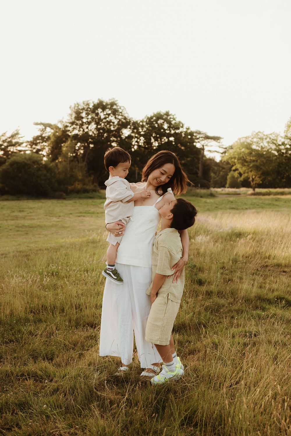 Three people in white and beige clothing embrace lovingly in a sunlit grassy field during golden hour.