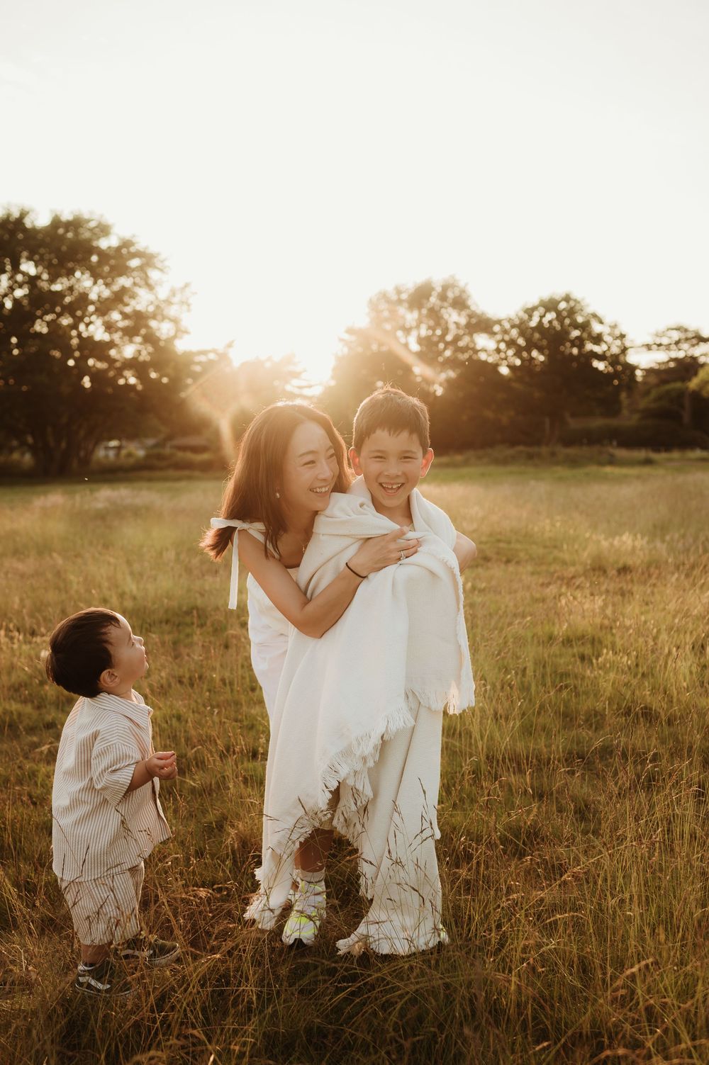 Three children in white outfits play together in a sunlit grassy field at golden hour.