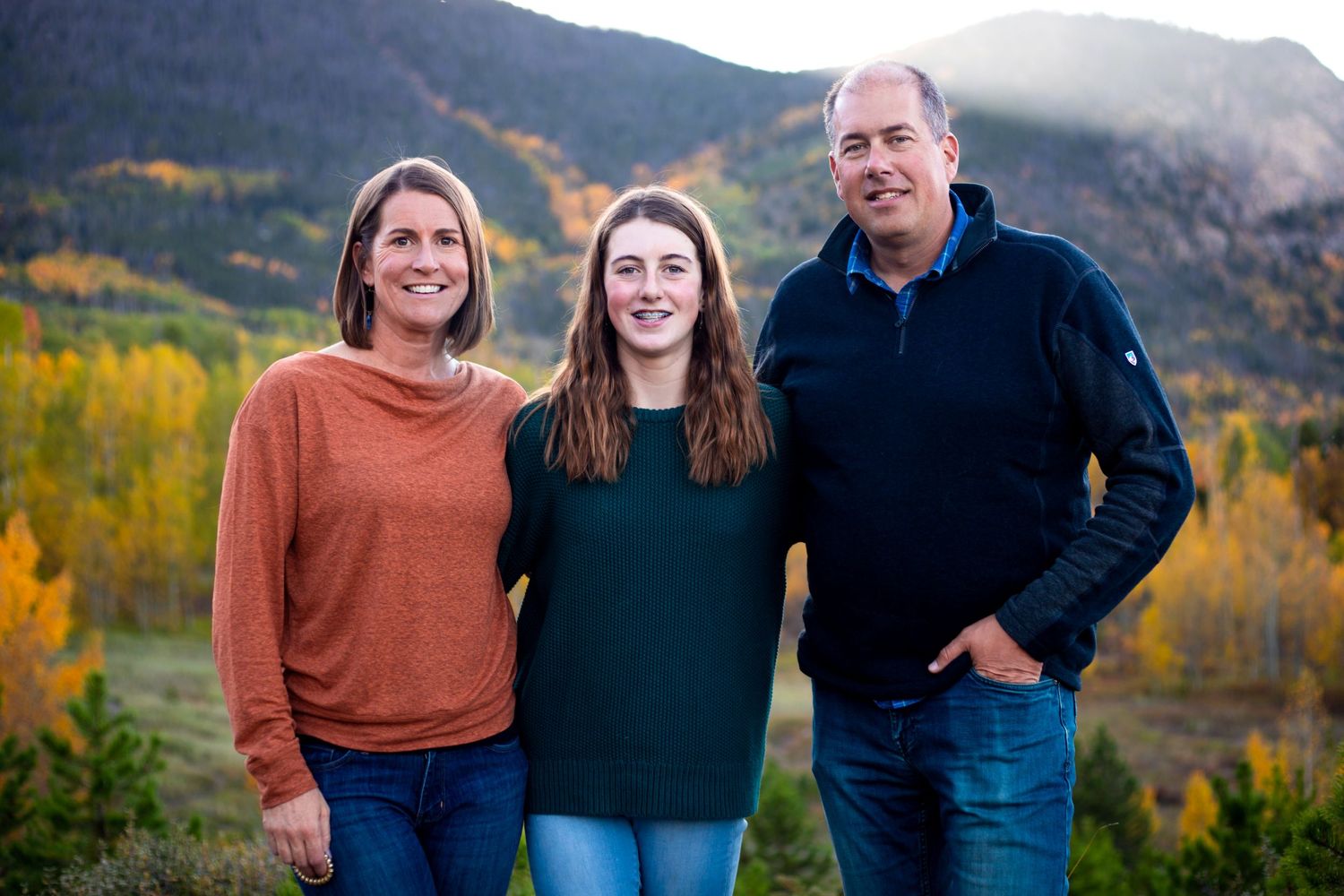 A family of three standing with their arms around each other smiling at the camera in the Rocky Mountains