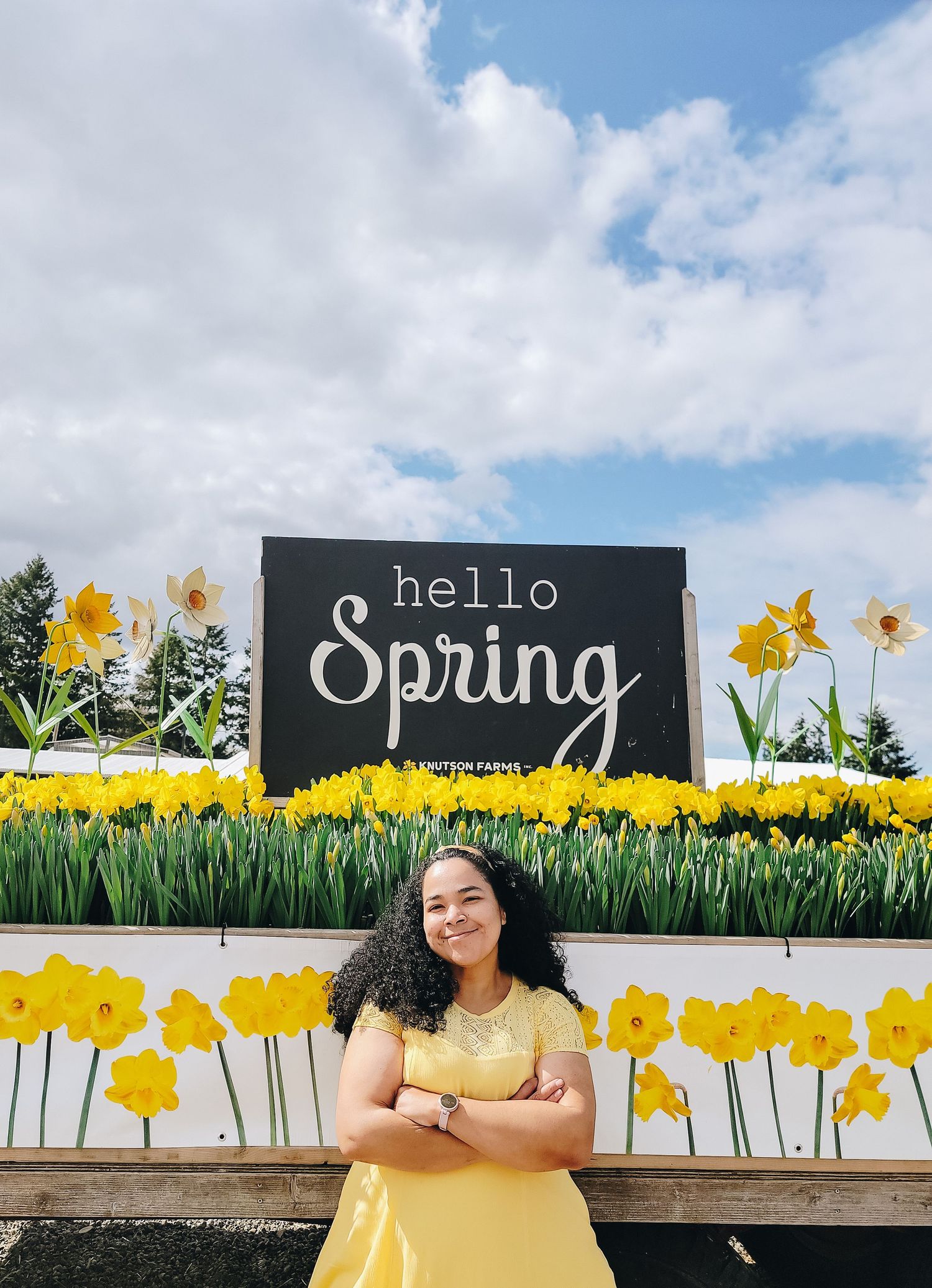 Woman posing in front of “Hello Spring” sign surrounded by daffodils at Knutson Farms