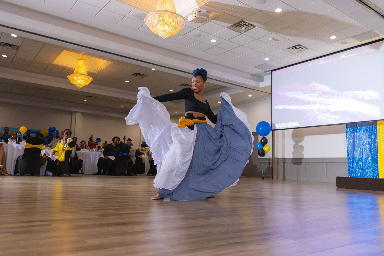 A dancer in a flowing blue and white dress performs a graceful twirl in a ballroom with crystal chandeliers overhead.