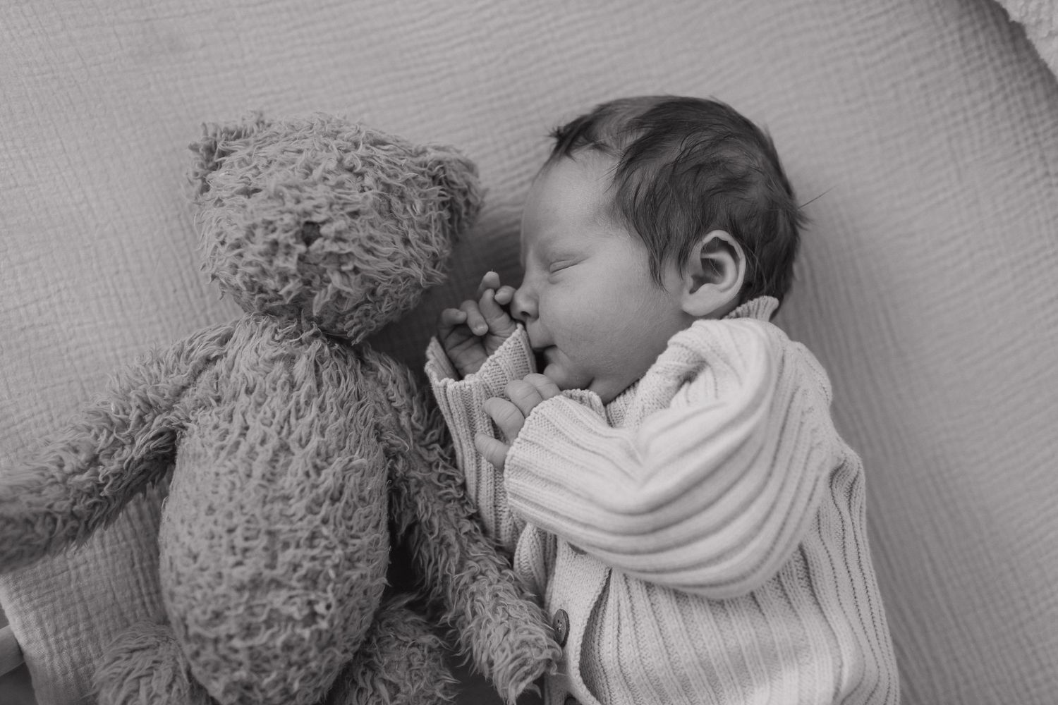 A sleeping baby lies next to a plush teddy bear on soft bedding in black and white.