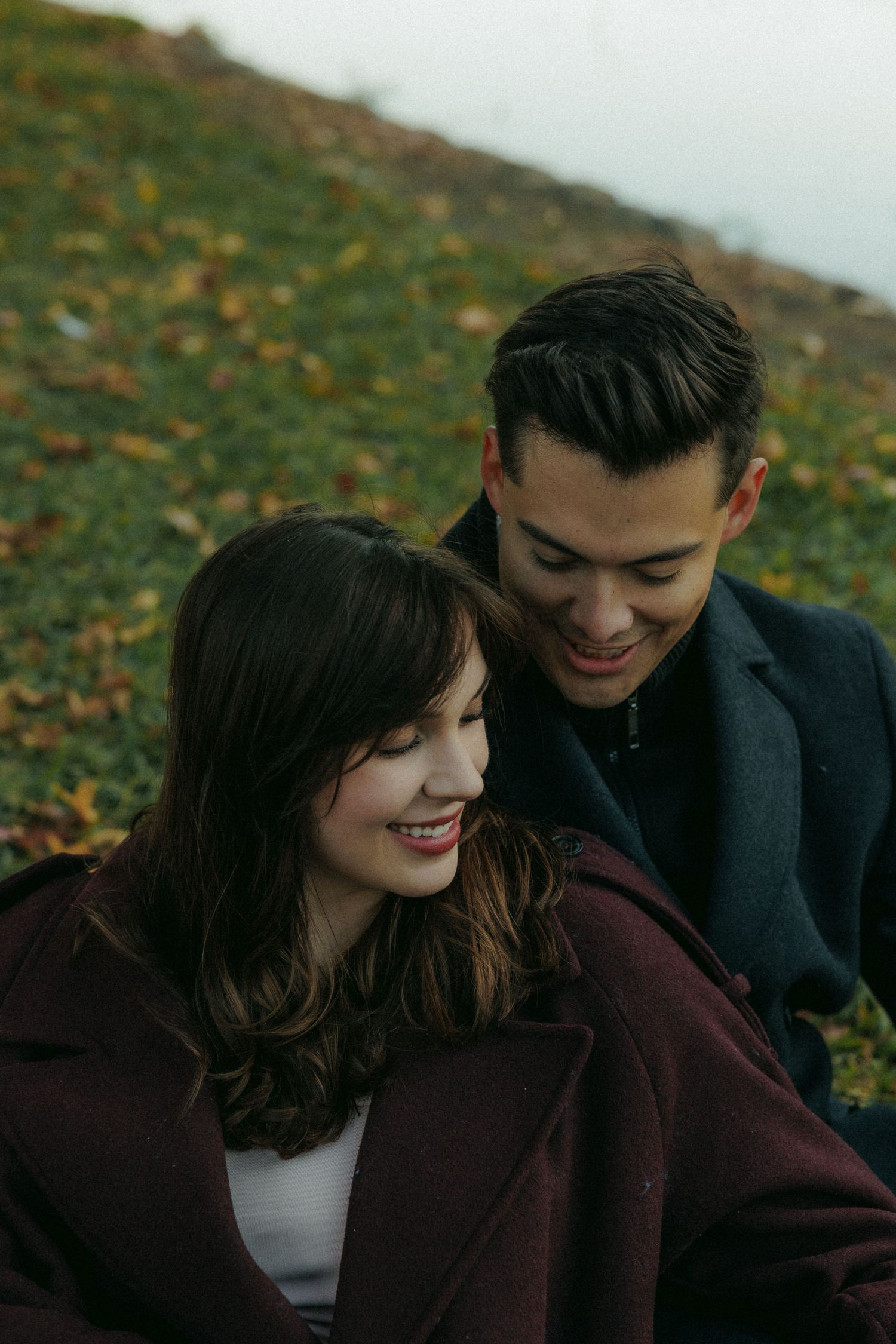 A couple sits close together on grass covered with autumn leaves in muted tones.