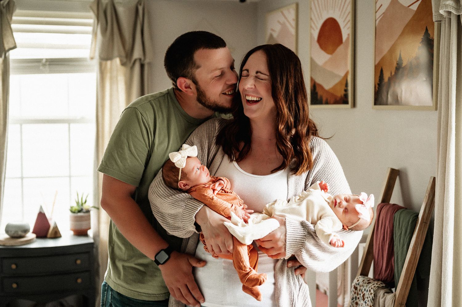 A joyful couple sharing a tender moment while holding newborn twins in a cozy bedroom setting. Virginia family photographer.