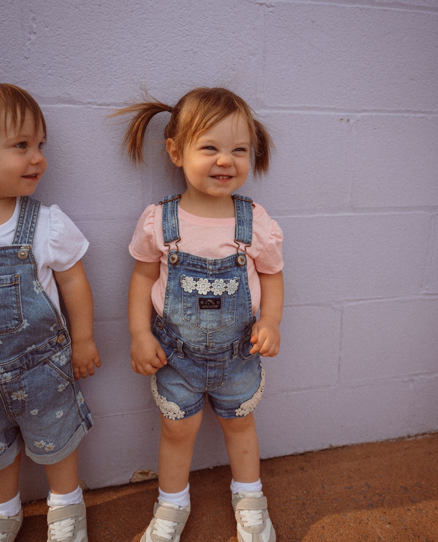 Smiling toddler girl with pigtails wearing denim overalls and pink shirt standing against a gray wall.
