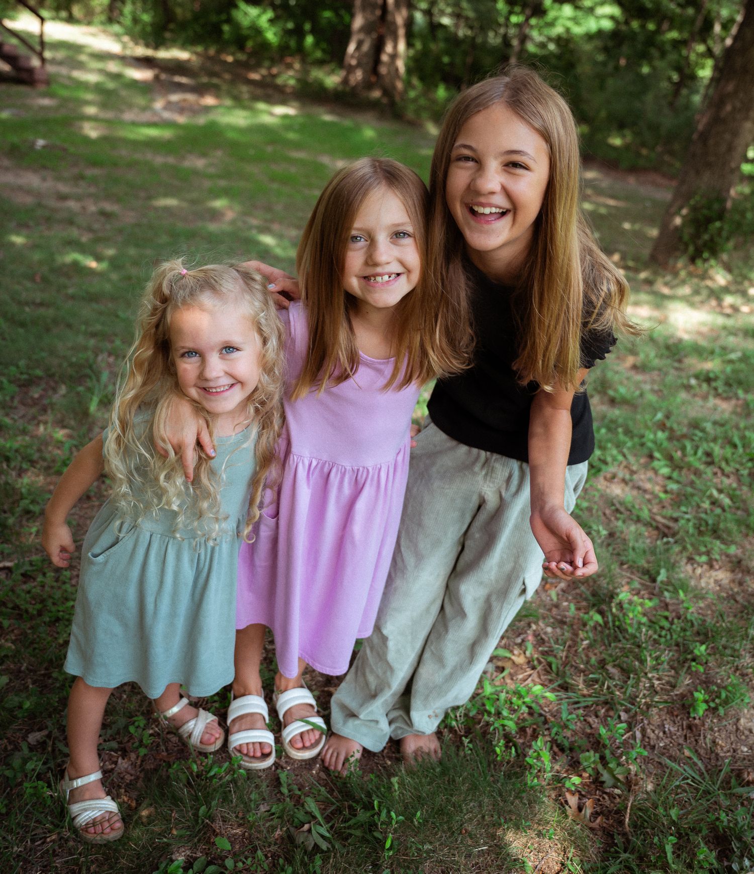 Three smiling girls pose together outdoors in a lush green park, wearing casual summer dresses and sandals.