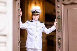 Someone in a crisp white naval dress uniform stands smiling between ornate wooden doors with decorative hardware.