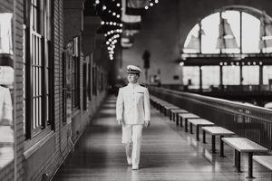 A solitary figure in white uniform walks through a long covered walkway with arched windows filled with ambient light.