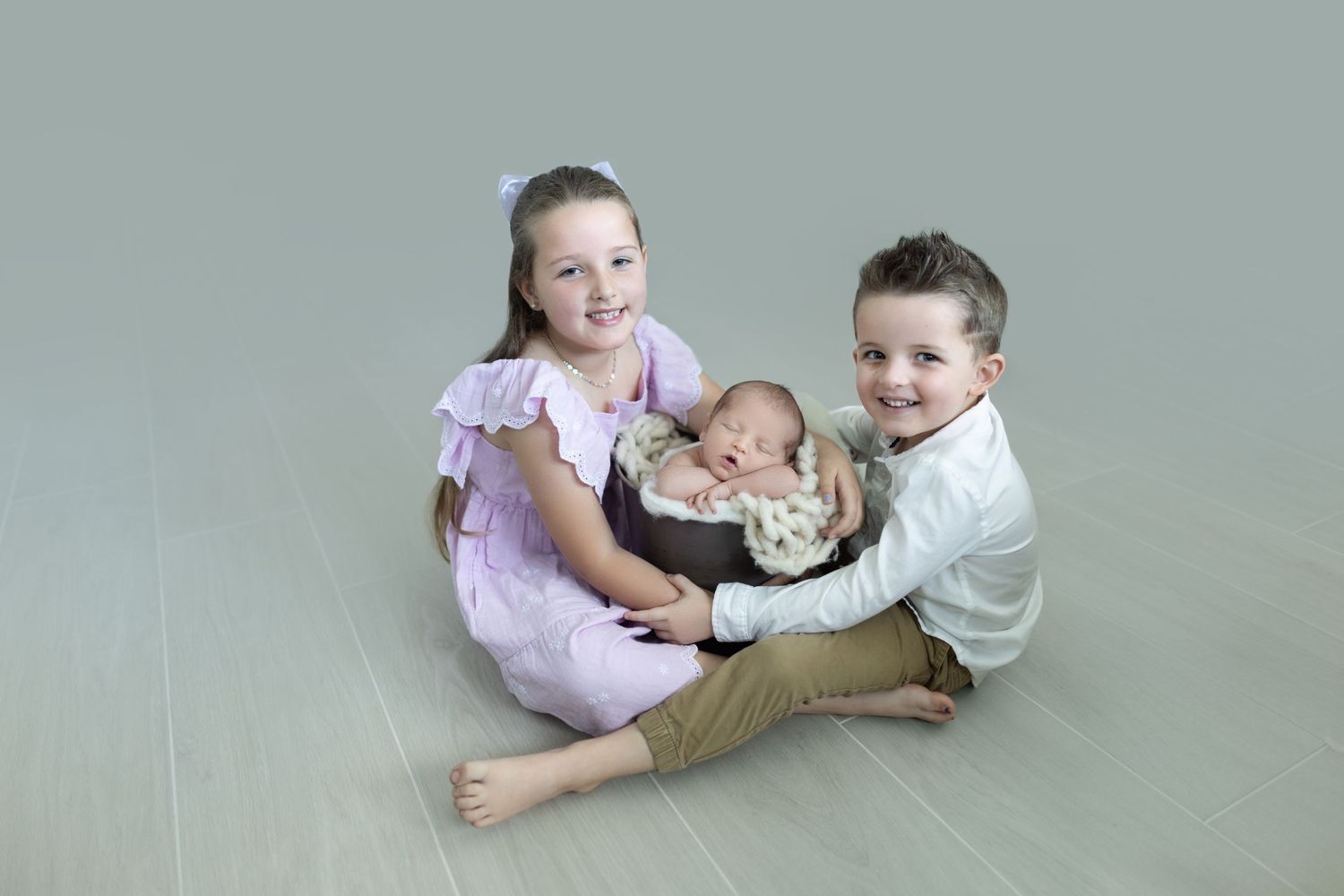 Three children sitting together on gray backdrop during studio photoshoot.
