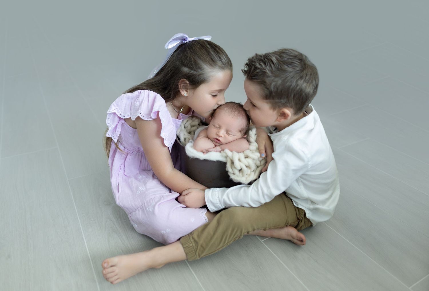 Young siblings sharing tender moment with baby on gray studio floor.