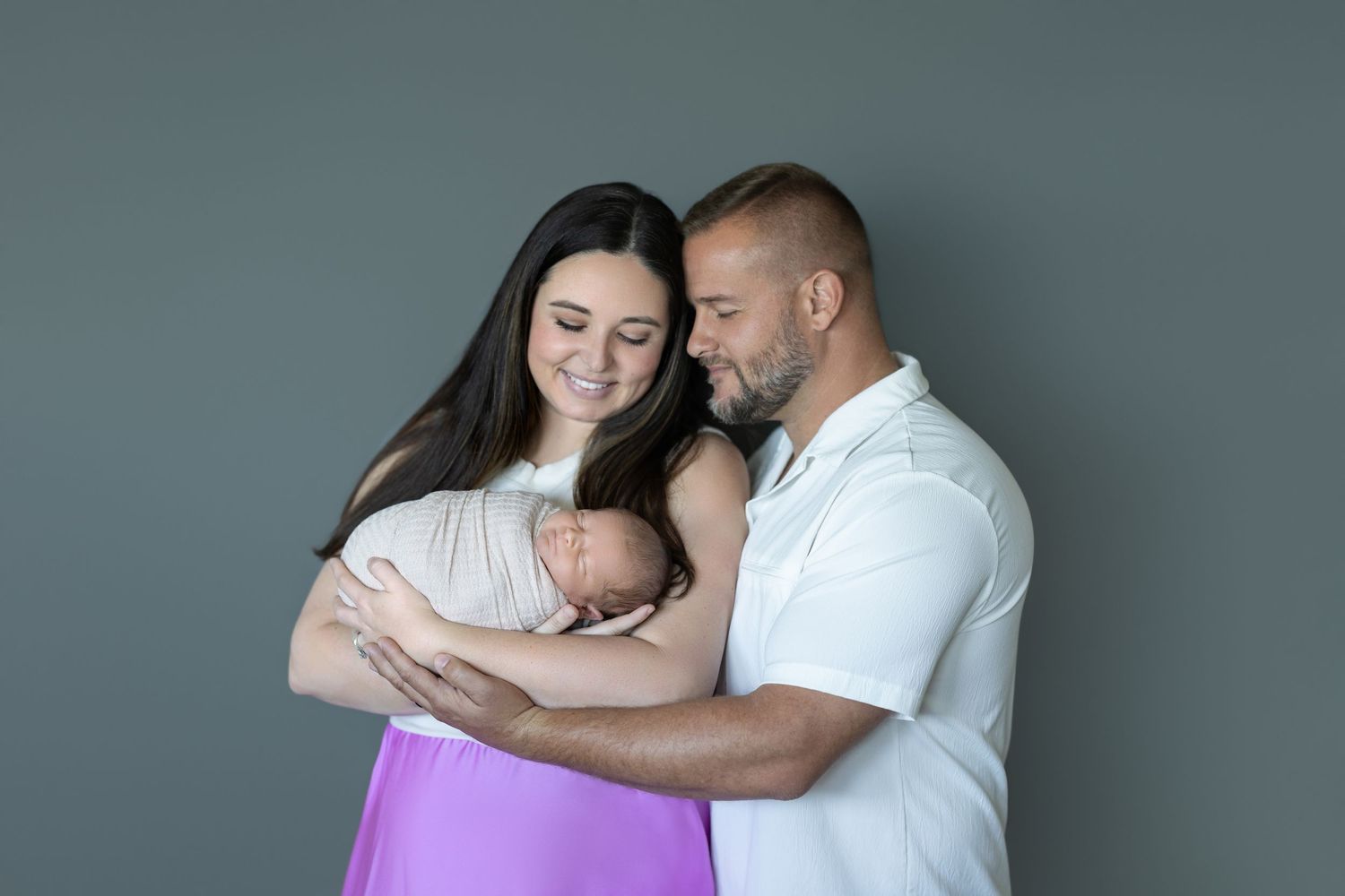 A tender family moment as parents embrace their newborn baby against a gray background.