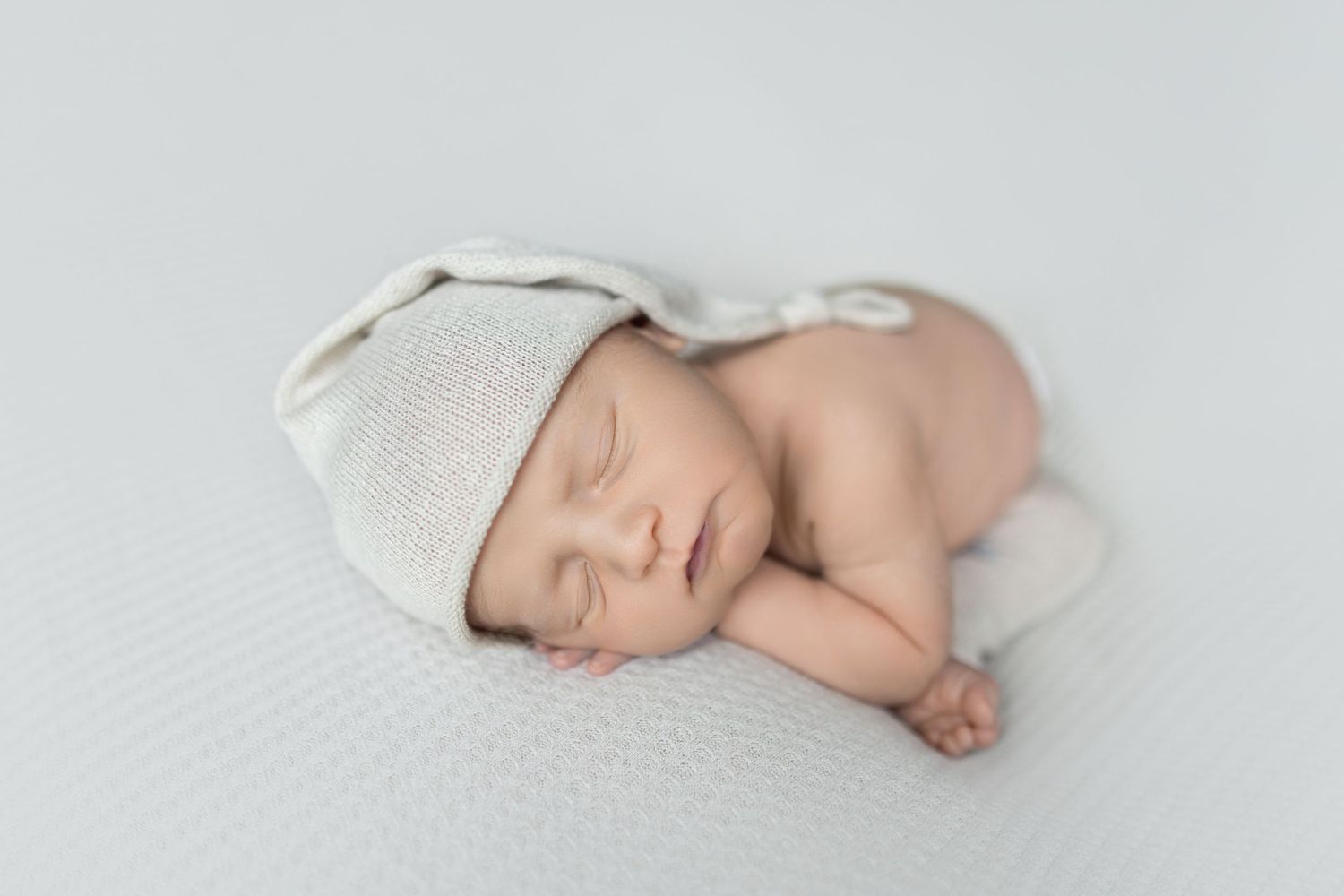 Sleeping newborn wearing white cap resting on textured white surface.