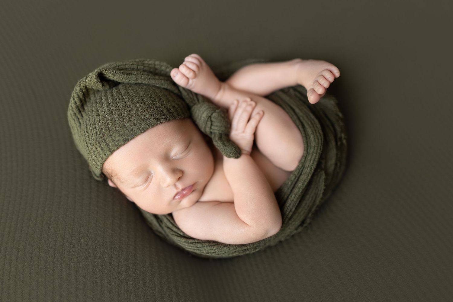 Sleeping newborn wrapped in olive green fabric on dark background.