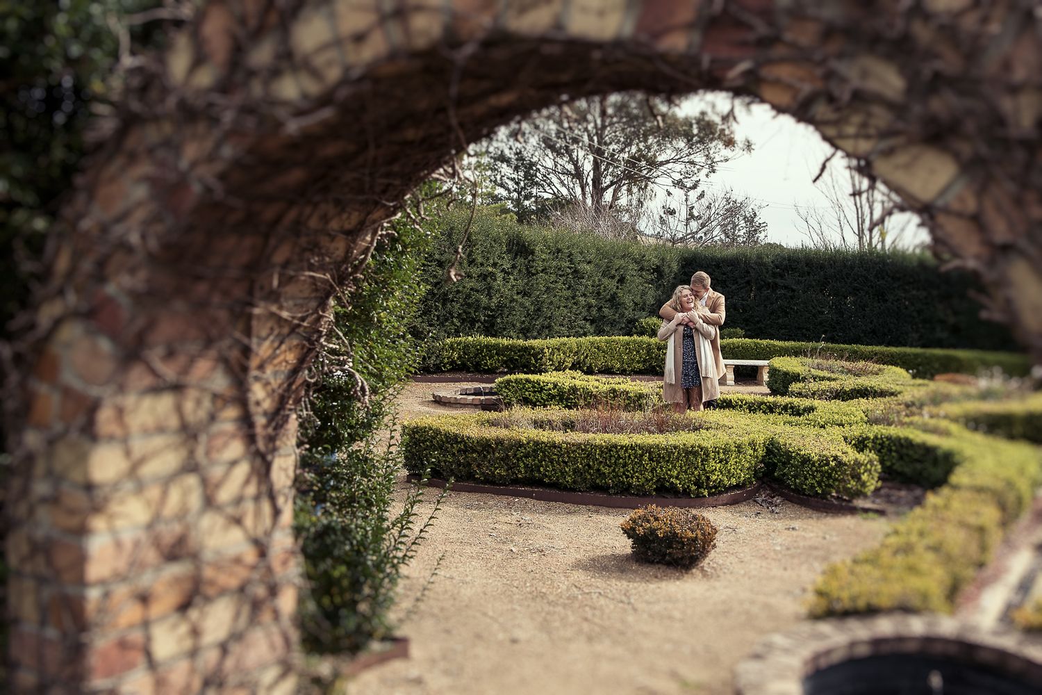 couple is embracing in a garden with neatly trimmed hedges, viewed through an archway.