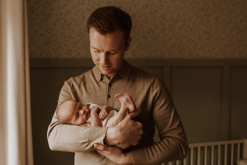 A tender moment is captured as a caregiver in a tan sweater gently holds a newborn baby in soft natural window light.