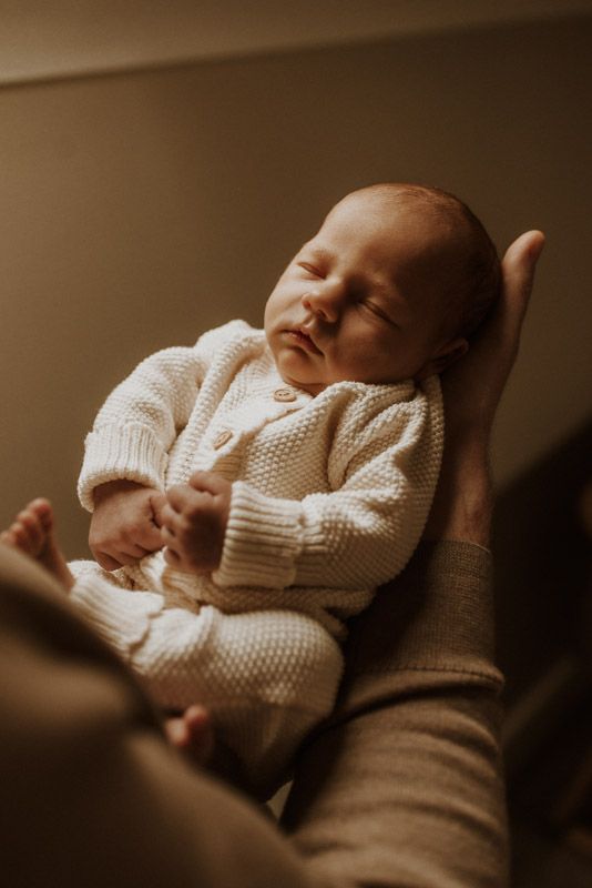 Sleeping newborn baby wearing cozy white knit sweater rests peacefully in caregiver's arms.
