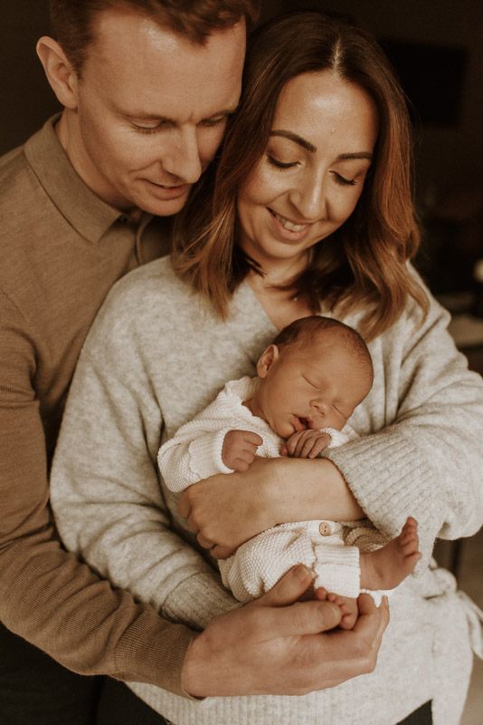 Couple in cozy sweaters holding and gazing lovingly at their sleeping newborn baby in warm, intimate lighting.