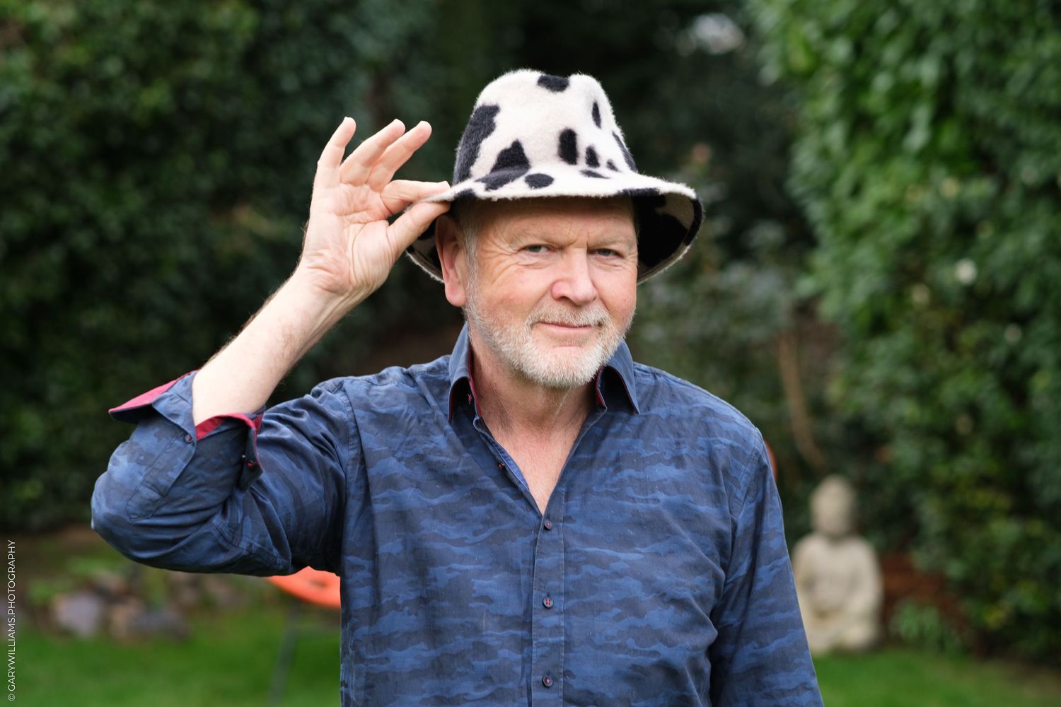 An older person in a denim shirt tips a panda-patterned hat while standing in front of green foliage.