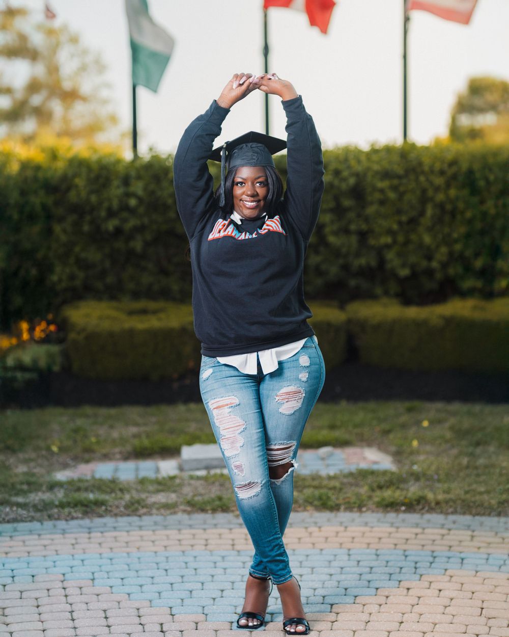 Smiling graduate in cap and sweatshirt poses with arms raised in ripped jeans outdoors near flags and hedges.