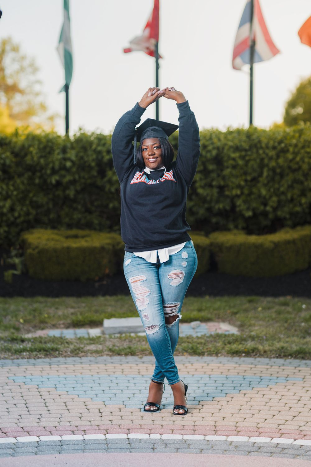 Young woman in graduation cap, ripped jeans, and heels posing with arms raised at outdoor campus plaza.