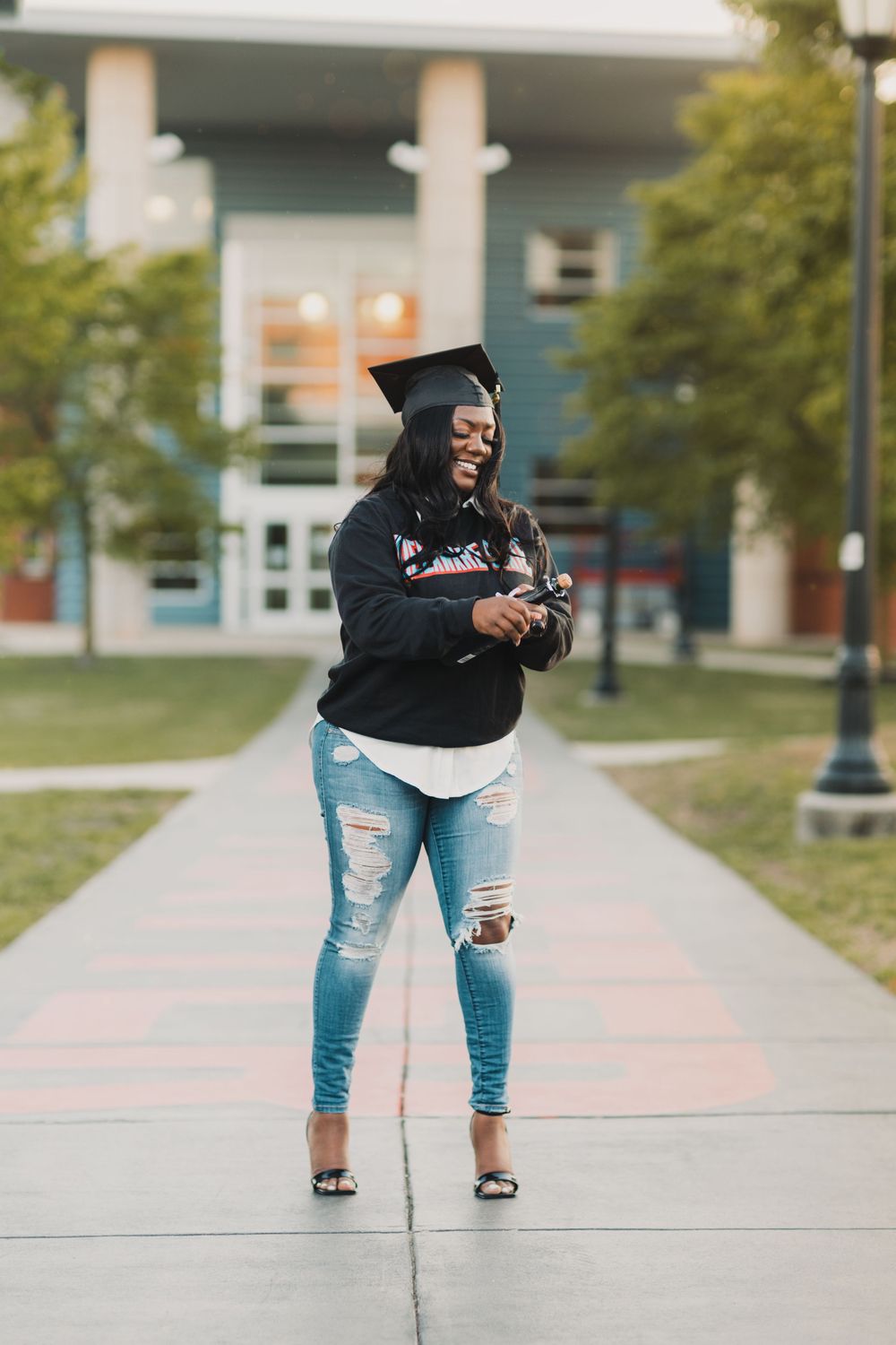 Graduate in cap and ripped jeans smiling at phone on college campus walkway.