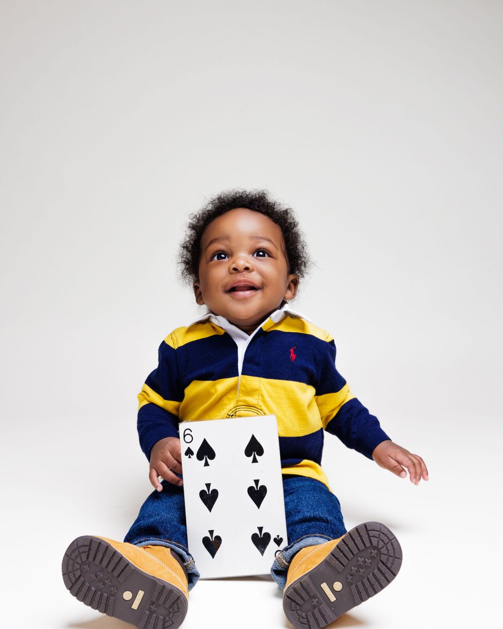 Baby in striped polo outfit sits happily with playing card against white background.