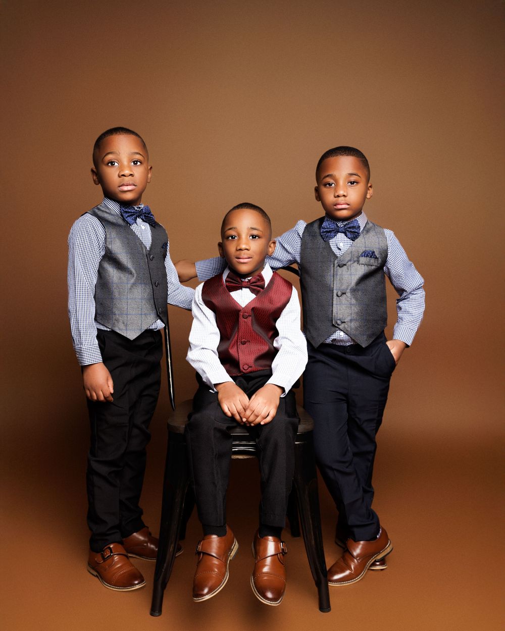 Three young boys in formal attire with vests and bowties pose for portrait against brown background.