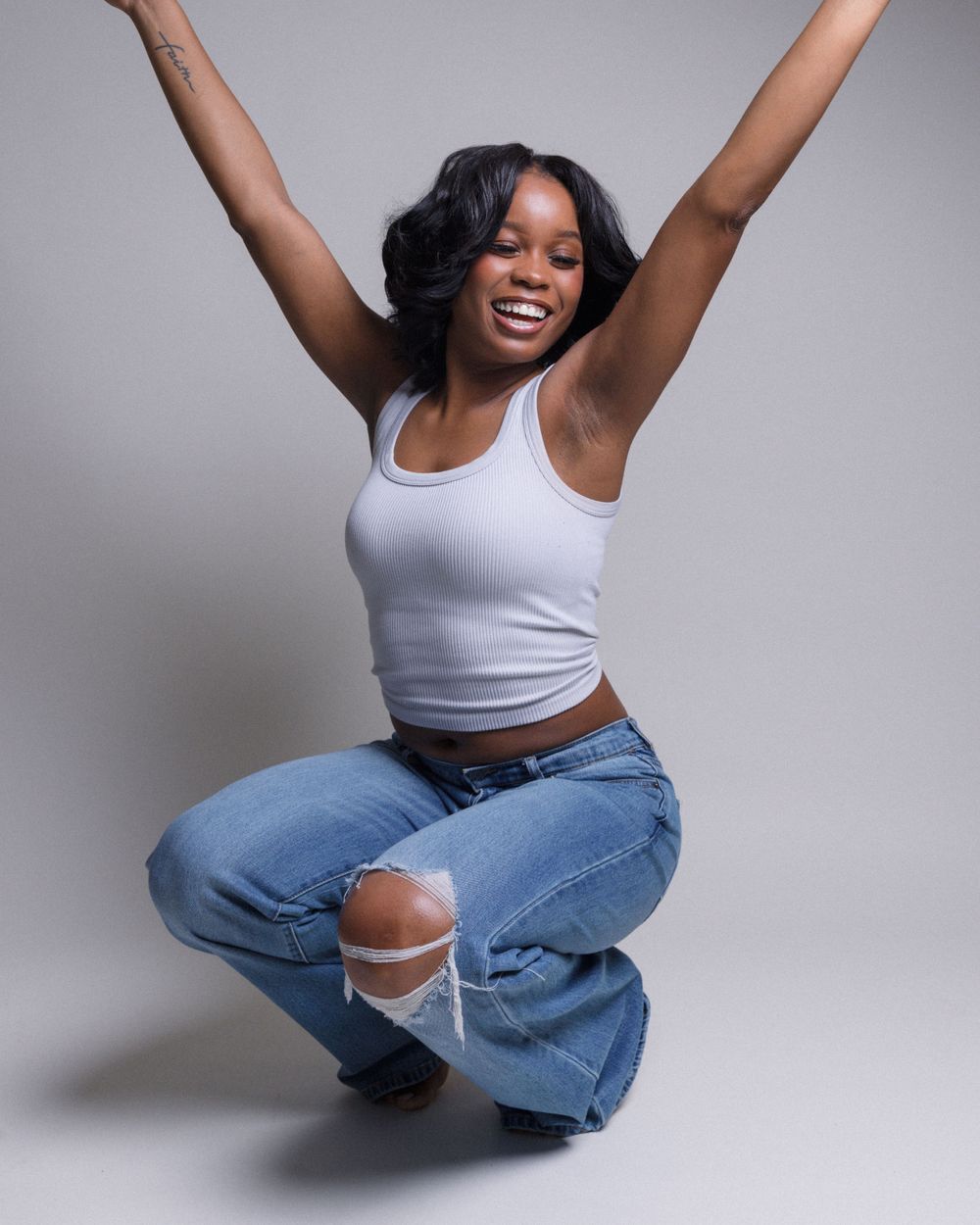 A joyful dancer in ripped jeans and white tank top strikes an energetic pose against a gray background.