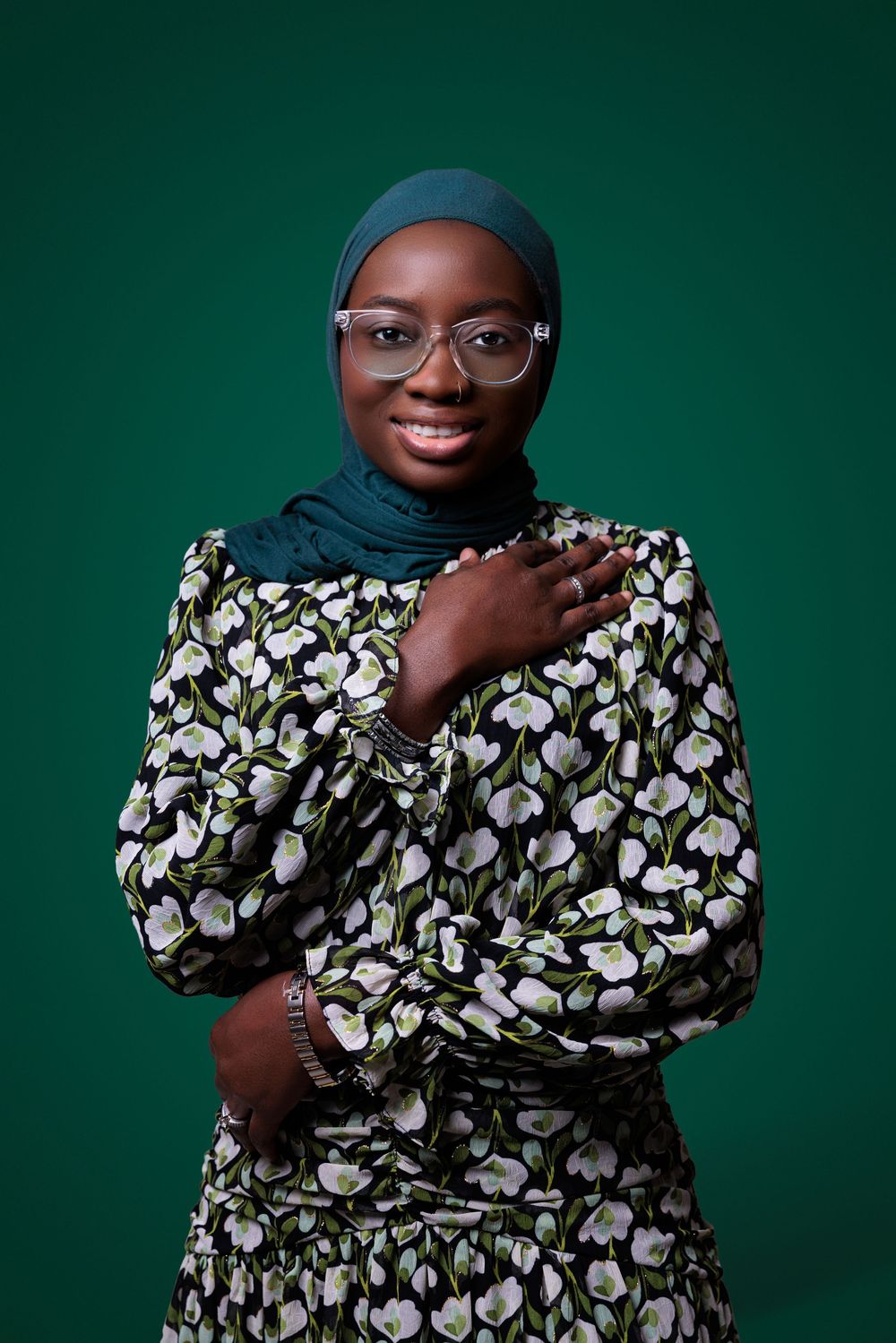 Smiling woman in floral dress and teal hijab with glasses, hand on chest, posed against green background.
