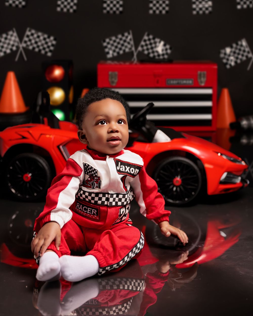Baby boy in red racing suit named Jaxon sits before toy race car and checkered flag backdrop.
