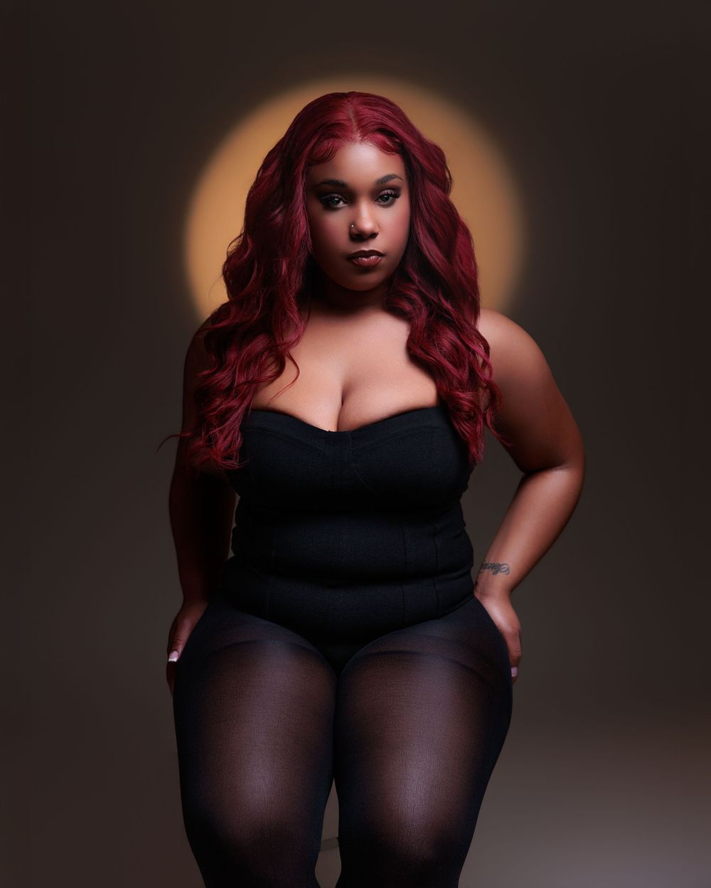 A plus-size woman with long red hair poses confidently in a black strapless top and leggings against a dark background.