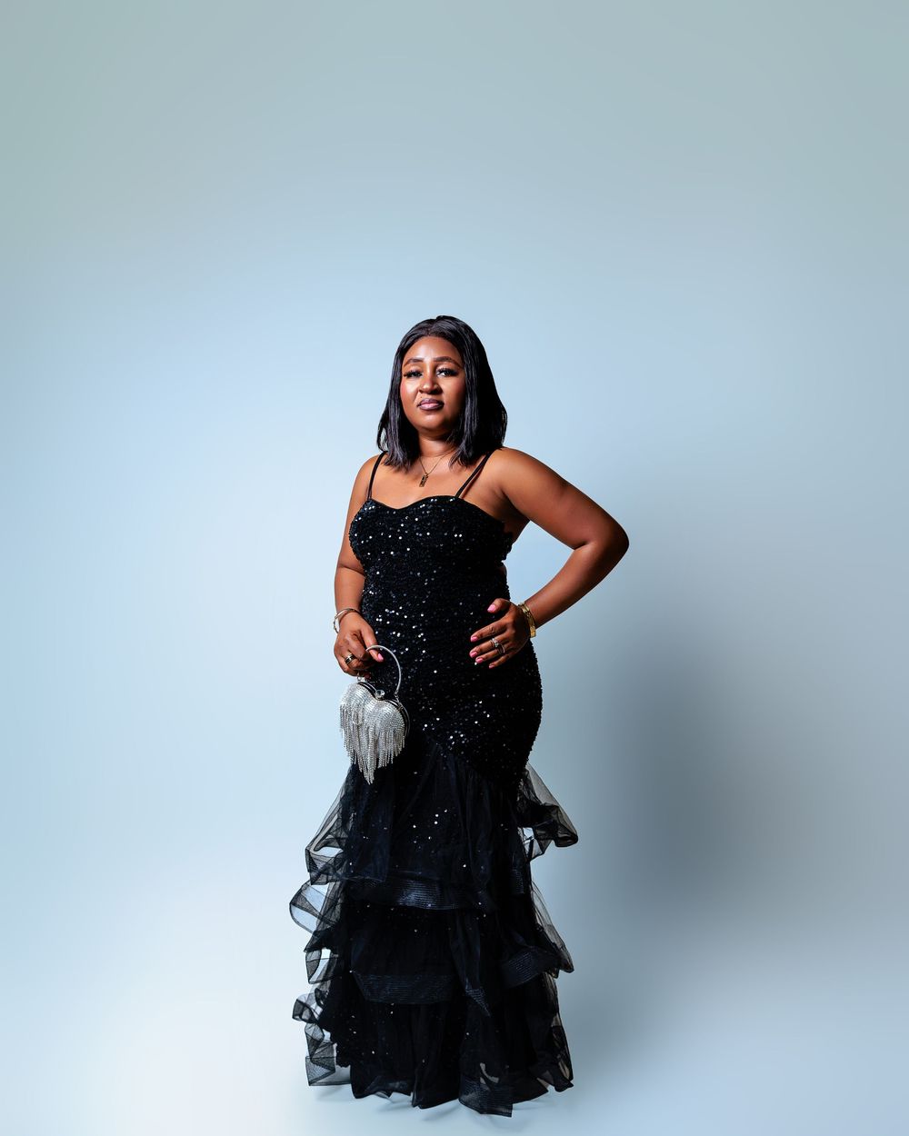 A woman poses in a black sequined mermaid gown with ruffled tulle hem, holding a silver clutch against a light backdrop.