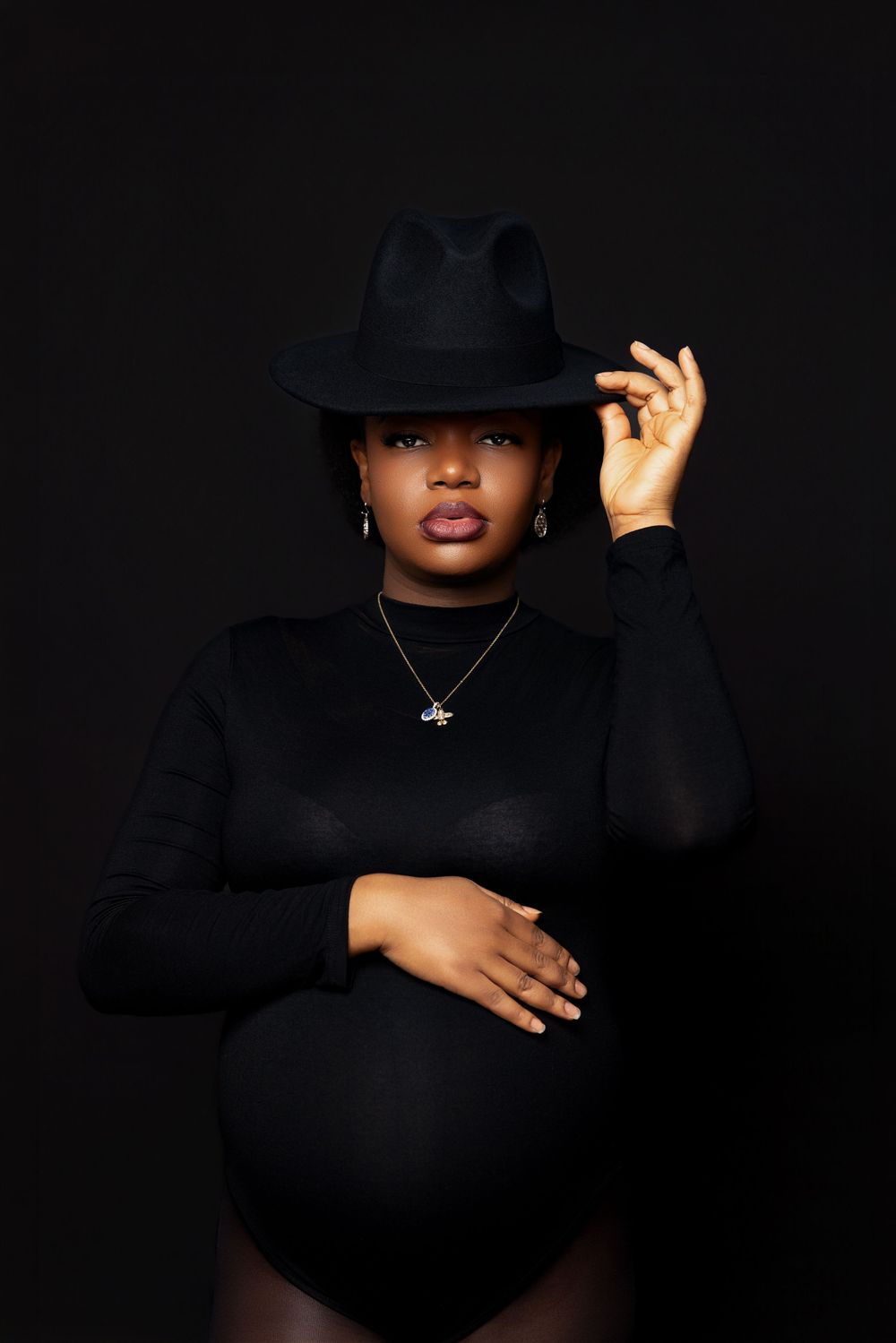 Pregnant Black woman in black bodysuit and fedora hat poses confidently against dark background.